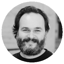 A bearded man with medium-length dark hair smiles at the camera in this black-and-white portrait of Chris Petridis, framed by a circular border.