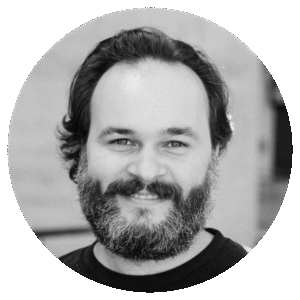A bearded man with medium-length dark hair smiles at the camera in this black-and-white portrait of Chris Petridis, framed by a circular border.