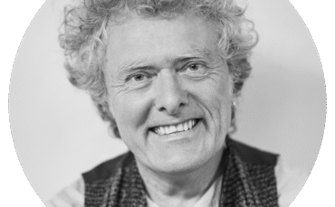 Smiling older person with curly hair, wearing a patterned vest over a light shirt, photographed in black and white with a plain background—Quincy Grant exudes warmth and character in this timeless portrait.