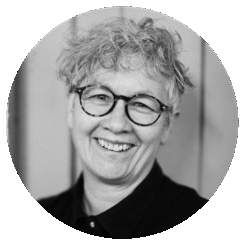 A person with short, curly gray hair and round glasses smiles warmly. They are wearing a dark top, and the background appears to be wooden panels. The photo is black and white and framed in a circular crop.