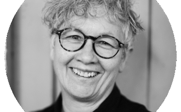 A person with short, curly gray hair and round glasses smiles warmly. They are wearing a dark top, and the background appears to be wooden panels. The photo is black and white and framed in a circular crop.