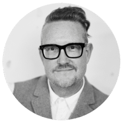 A person with short, side-shaved hair and glasses is wearing a suit jacket and collared shirt, smiling slightly while looking at the camera. The photo is black and white and framed in a circular border.