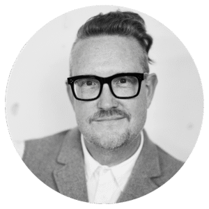 A person with short, side-shaved hair and glasses is wearing a suit jacket and collared shirt, smiling slightly while looking at the camera. The photo is black and white and framed in a circular border.