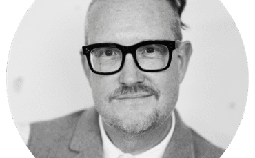 A person with short, side-shaved hair and glasses is wearing a suit jacket and collared shirt, smiling slightly while looking at the camera. The photo is black and white and framed in a circular border.