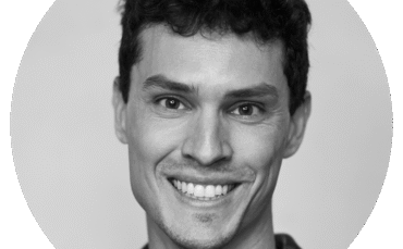 A black and white portrait of Darian Tregenza, a smiling young man with short, curly hair, wearing a collared shirt, facing the camera against a plain background.