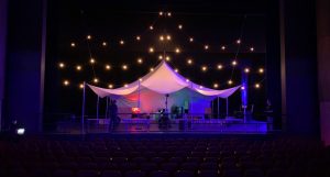 A tent, illuminated by strings of lights, sits on a theatre stage. Technicians, dressed in black, are at work.