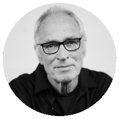 Black and white portrait of David McLean, an older man with short, light hair, wearing glasses and a dark shirt, looking directly at the camera with a slight smile. The background is plain and uncluttered.