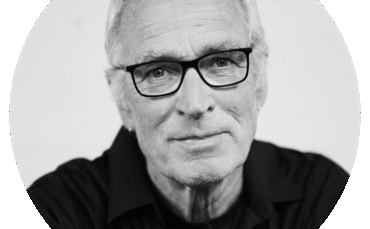 Black and white portrait of David McLean, an older man with short, light hair, wearing glasses and a dark shirt, looking directly at the camera with a slight smile. The background is plain and uncluttered.