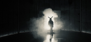 A figure wearing a costume and mask with two canisters attached stands in a doorway surrounded by fog. The scene is dimly lit, creating a mysterious atmosphere with shadows and soft lighting. The background is blurred, emphasizing the silhouetted figure.