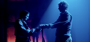 Two people stand facing each other in a dimly lit, blue-hued theatre space. They hold a scarf together between them. Shadows cast behind them create a dramatic atmosphere.
