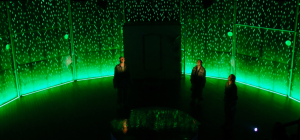 A dimly lit stage features three individuals standing apart, surrounded by tall, curved walls with a glowing green pattern resembling rain. The floor reflects the green light, creating an immersive, futuristic atmosphere.
