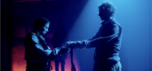 Two people stand facing each other in a dimly lit, blue-hued theatre space. They hold a scarf together between them. Shadows cast behind them create a dramatic atmosphere.