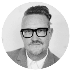 A man with short, slicked-back hair on top and shaved sides, wearing black glasses, a mustache, and a light-colored blazer over a white shirt, smiles slightly at the camera. The image is black and white.