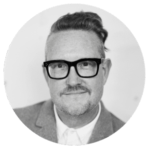 A man with short, slicked-back hair on top and shaved sides, wearing black glasses, a mustache, and a light-colored blazer over a white shirt, smiles slightly at the camera. The image is black and white.