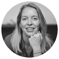 Black and white portrait of a smiling woman with light hair, resting her chin on her fist. She wears a ring and bracelet and looks directly at the camera, set against a blurred background inside a circular frame.