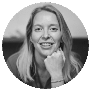 Black and white portrait of a smiling woman with light hair, resting her chin on her fist. She wears a ring and bracelet and looks directly at the camera, set against a blurred background inside a circular frame.