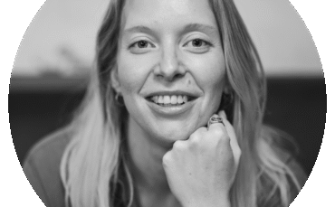 Black and white portrait of a smiling woman with light hair, resting her chin on her fist. She wears a ring and bracelet and looks directly at the camera, set against a blurred background inside a circular frame.