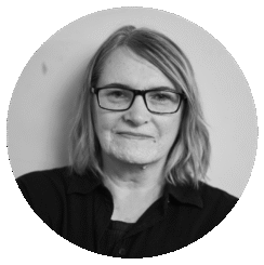 A woman with shoulder-length hair wearing glasses and a dark top looks directly at the camera with a slight smile, posed against a plain light background.