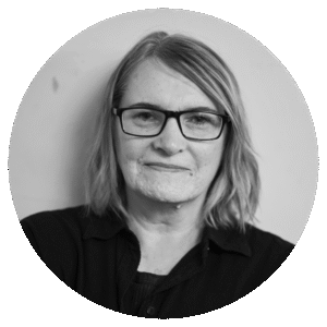 A woman with shoulder-length hair wearing glasses and a dark top looks directly at the camera with a slight smile, posed against a plain light background.