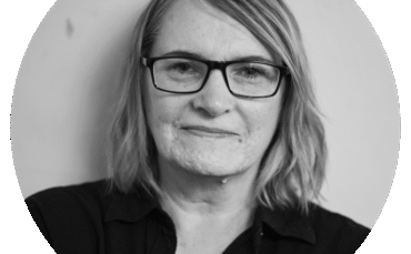 A woman with shoulder-length hair wearing glasses and a dark top looks directly at the camera with a slight smile, posed against a plain light background.
