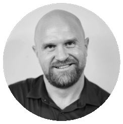 A bald man with a beard, wearing a dark collared shirt, smiles at the camera. The photo is black and white and set within a circular frame.