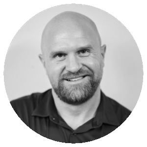 A bald man with a beard, wearing a dark collared shirt, smiles at the camera. The photo is black and white and set within a circular frame.