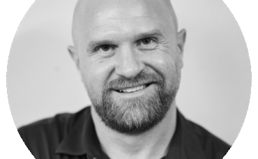 A bald man with a beard, wearing a dark collared shirt, smiles at the camera. The photo is black and white and set within a circular frame.