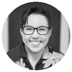Black and white portrait of Jess Wolfendale, a person with short dark hair, glasses, and a floral shirt, smiling broadly at the camera.