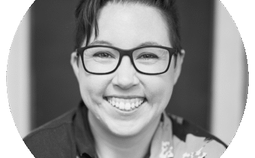 Black and white portrait of Jess Wolfendale, a person with short dark hair, glasses, and a floral shirt, smiling broadly at the camera.