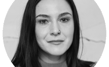 Black and white portrait of a woman with long dark hair, a nose ring, and a slight smile, looking directly at the camera, set against a plain light background.