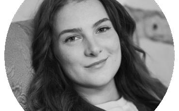 Ren Williams, a young woman with long, wavy hair, smiles softly while sitting on a patterned chair. The black and white image is framed in a circular crop.