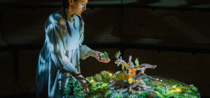 A performer costumed in a Victorian-period dress, with braided hair over her shoulders, leans over a diorama of a garden. The garden is lit with tiny lights, bringing a magical feel to the textured green miniature landscape. She is holding leaves in her hand which appear to have been plucked from a small tree at the centre of the garden.