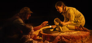 Three people in costume kneel around a round table with a soil-filled tray. A tiny illuminated snail adds to the tableau, as one person uses a doll to interact with the scene.