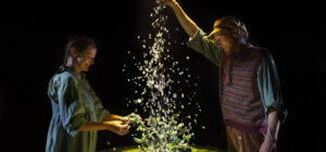 Two people stand around a round table covered in greenery and small plants. One pours a stream of white particles, possibly petals or confetti, as both smile and interact with the miniature landscape, illuminated by dramatic theatre lighting.