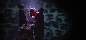 Three people in period costumes dramatically pose under a spotlight against a curtain, showing artwork resembling a stone wall. The group are animating a puppet so that it appears to be climbing the wall.