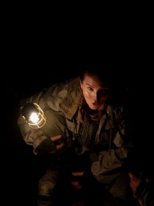 Person in a dark setting, partially illuminated by a hanging light, wearing a heavy jacket and looking upwards with a surprised expression.