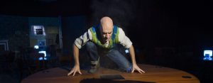 An actor kneels on a wooden floor, illuminated under a spotlight, casting a shadow resembling a wolf onto the table's surface.