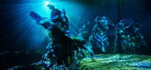 Performers in animal-like costumes dance under dramatic blue and green stage lighting, as their movements cast dynamic shadows and patterns across the floor and ceiling in a mysterious, immersive theatre setting.