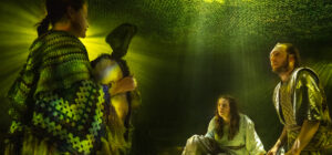 Three actors perform in scene, lit mysteriously by shafts of yellow-green light piercing through a net-like canopy. One performer, wearing a knitted poncho, stands as the others sit nearby, their faces tense and emotional.