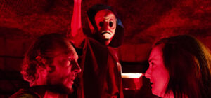 Two people stand face-to-face in dramatic red lighting, while a third, cloaked and masked, raises a card behind them, creating a tense and mysterious atmosphere.