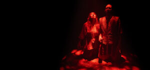 Two people in medieval-style clothing kneel close together, looking upward, bathed in dramatic red lighting with darkness surrounding them on stage.