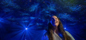 A woman smiles while standing under an artistic blue and green light installation, with netting overhead and bright spotlights shining in the background.
