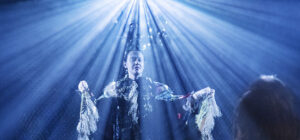 A person in dramatic, fringed clothing stands illuminated by bright blue rays of light, as they face an onlooker who has their back to the camera in a theatrical setting.