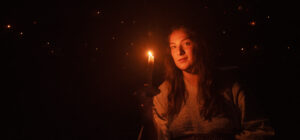 A young woman with long hair holds a lit candle in a dark room, softly illuminating her face. Small, glowing lights are scattered in the background, evoking a mysterious feeling.