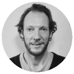 Black and white portrait of Nathan O'Keefe, a man with short, wavy hair and a beard, wearing a casual shirt and sweater. He looks directly at the camera with a slight smile; the background is plain and softly blurred.