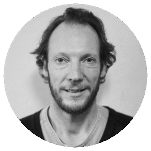Black and white portrait of Nathan O'Keefe, a man with short, wavy hair and a beard, wearing a casual shirt and sweater. He looks directly at the camera with a slight smile; the background is plain and softly blurred.