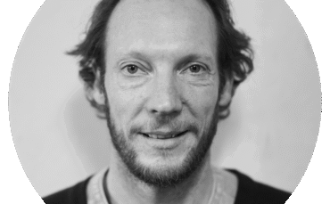 Black and white portrait of Nathan O'Keefe, a man with short, wavy hair and a beard, wearing a casual shirt and sweater. He looks directly at the camera with a slight smile; the background is plain and softly blurred.