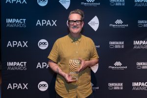 A person wearing glasses and a mustard-colored shirt smiles and holds a PAC Australia IMPACT Award trophy in front of a backdrop with logos for APAX, Impact Awards, Slingsby, and various sponsors.