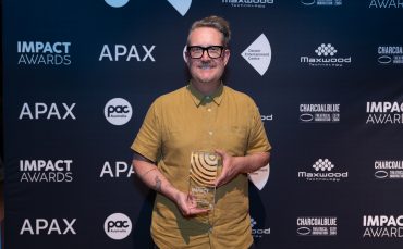 A person wearing glasses and a mustard-colored shirt smiles and holds a PAC Australia IMPACT Award trophy in front of a backdrop with logos for APAX, Impact Awards, Slingsby, and various sponsors.