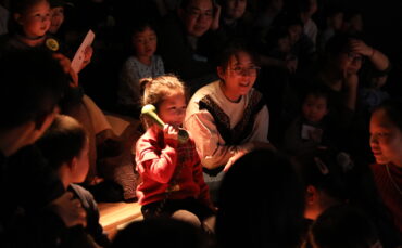 A young girl in a red sweater holds a green phone receiver to her ear, surrounded by adults and children sitting closely together in a dimly lit room, all watching something attentively.